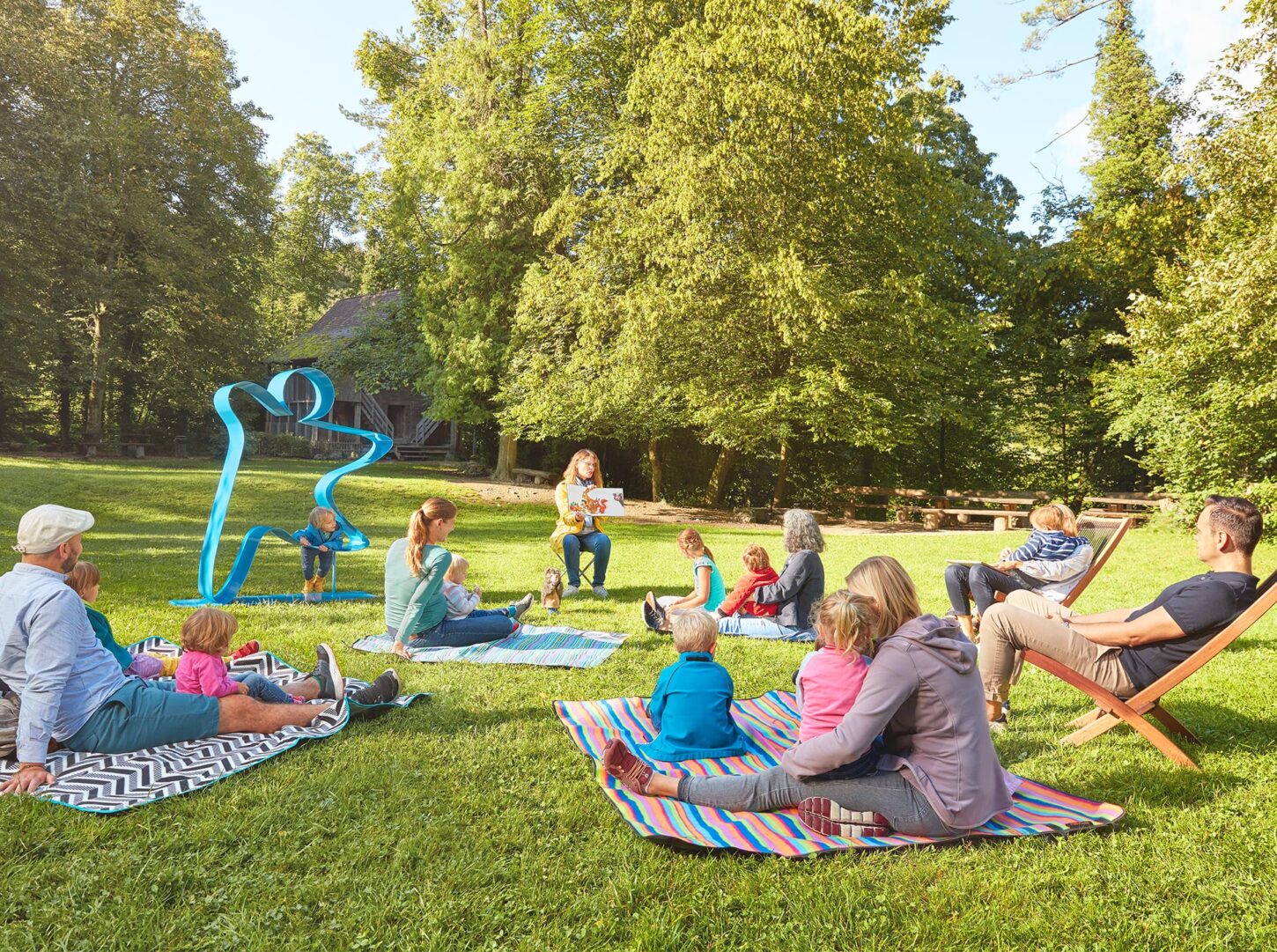 Auf einer grünen Wiese im Park sitzt eine Gruppe junger Eltern mit Kinder. Vor ihnen sitzt eine Lehrerin und erzählt eine Geschichte aus einem Buch. Ein kleiner Junge klettert auf dem menschengrossen blauen Stiftungslogo aus Metall herum, das links neben der Gruppe auf der Wiese steht.